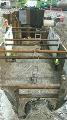Workers with a ladder in a steel-braced excavation, with a large pipe opening and a screened structure in the background