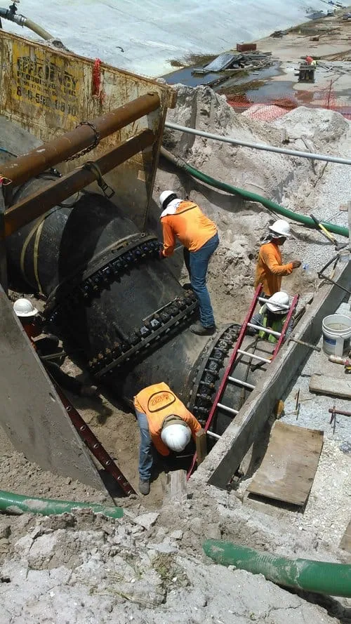 Workers guiding a large bolted flange pipe fitting into place with an excavator bucket, in a braced excavation