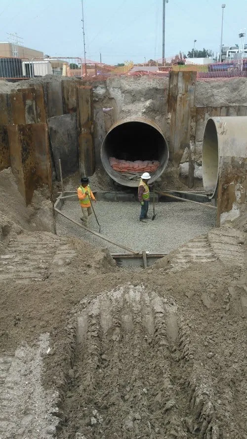Workers standing in a large steel-braced excavation between two large drainage pipe openings