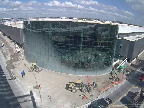 Curved glass terminal building exterior under construction with equipment on the surrounding plaza