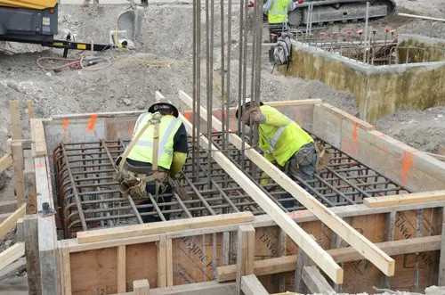 Two workers in safety vests tying rebar inside a wooden footing or pile-cap formwork frame