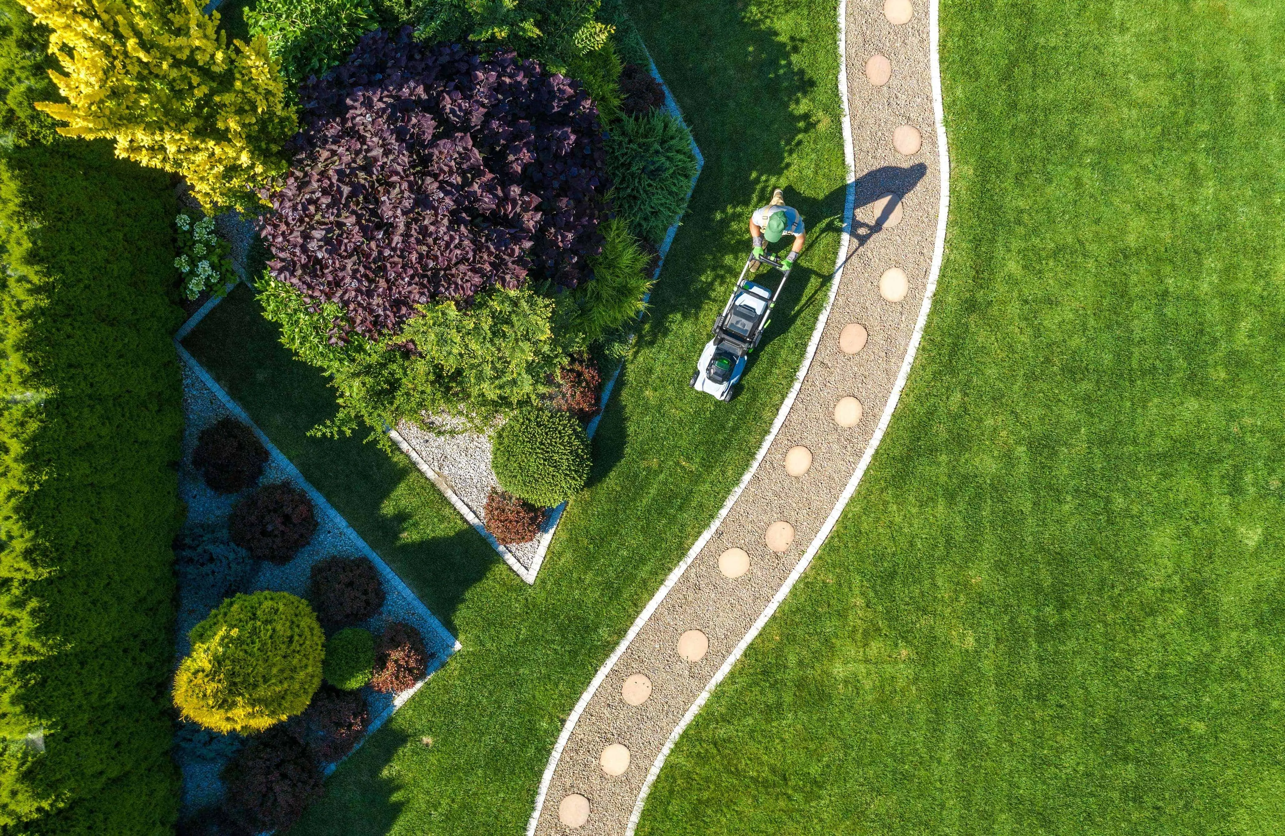 Landscaper mowing a lawn beside a curved paver path and planted garden beds
