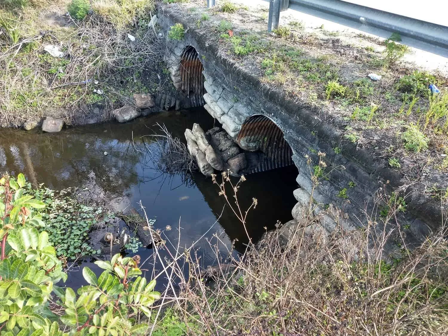 Deteriorated corrugated metal culvert pipes under a roadway, partially submerged in a canal