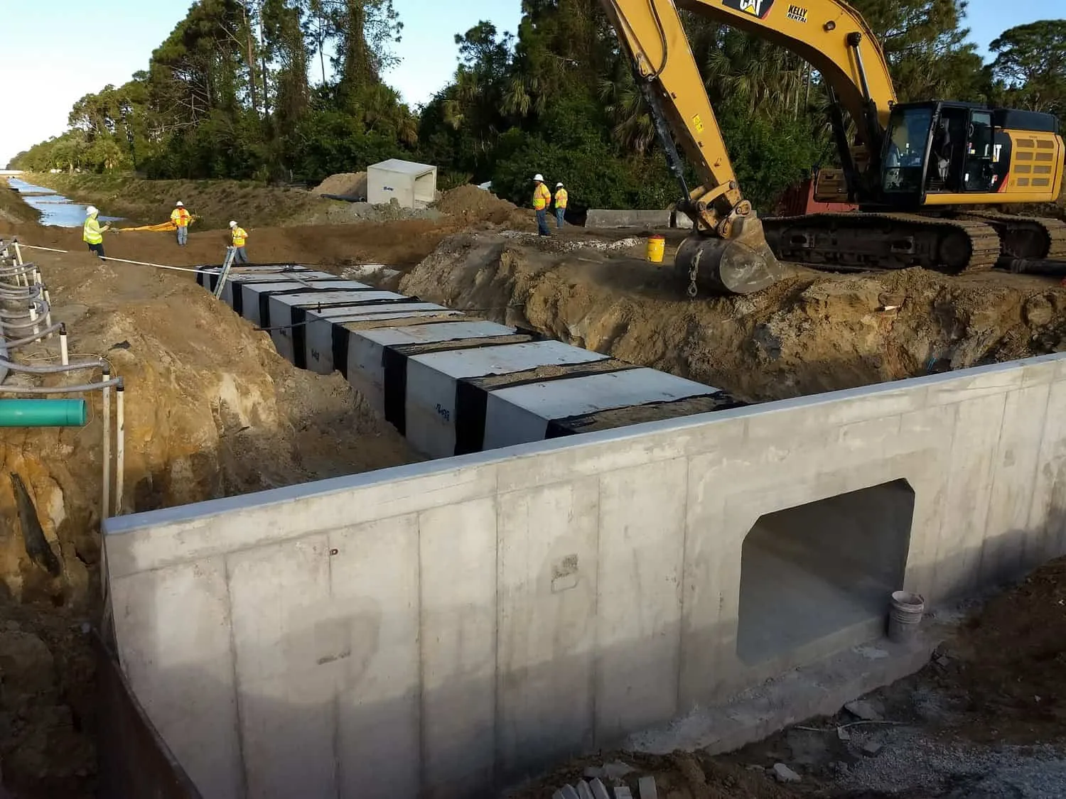 Excavator beside precast concrete box culvert sections and a finished culvert headwall along a canal