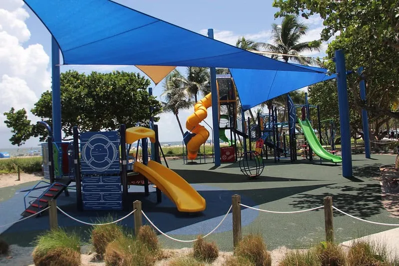 Shaded playground with slides and climbing equipment on a sand surface near a beach