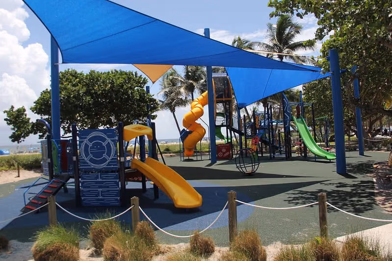 Shaded playground with slides and climbing equipment on a sand surface near a beach
