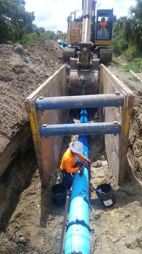 Worker in a hard hat connecting sections of blue water pipe in a supported trench, with an excavator above