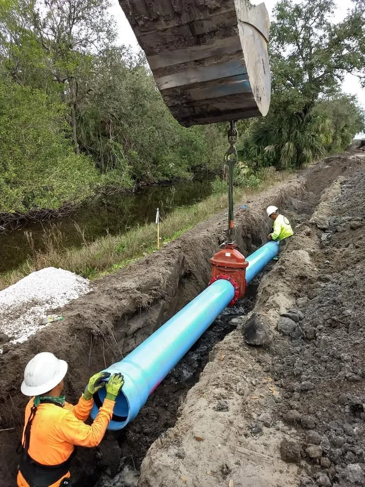 Excavator lowering a large blue water pipe and valve assembly into an open trench beside a canal, guided by two workers