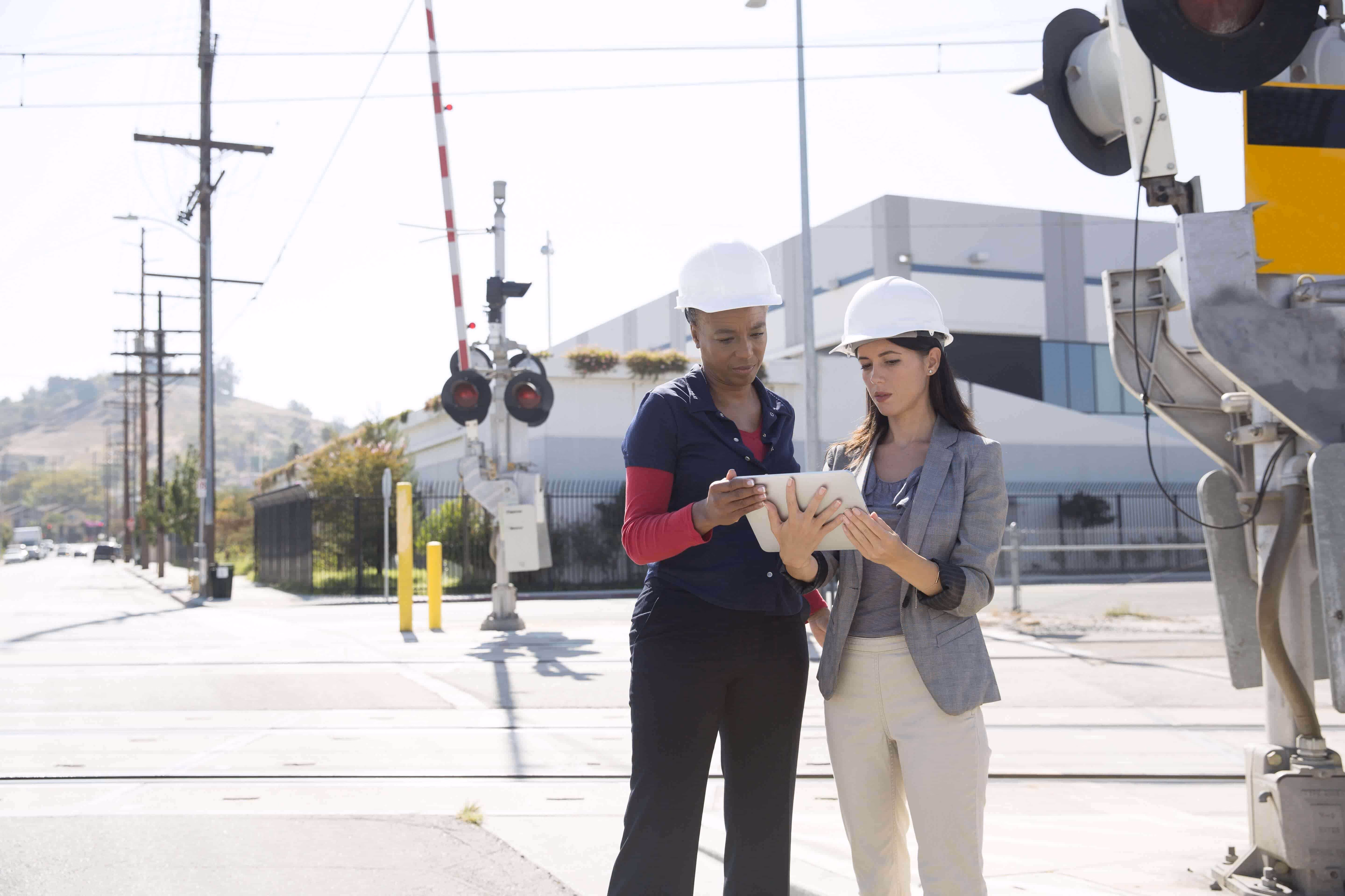 Two engineers in hard hats reviewing plans on a tablet at a railroad crossing