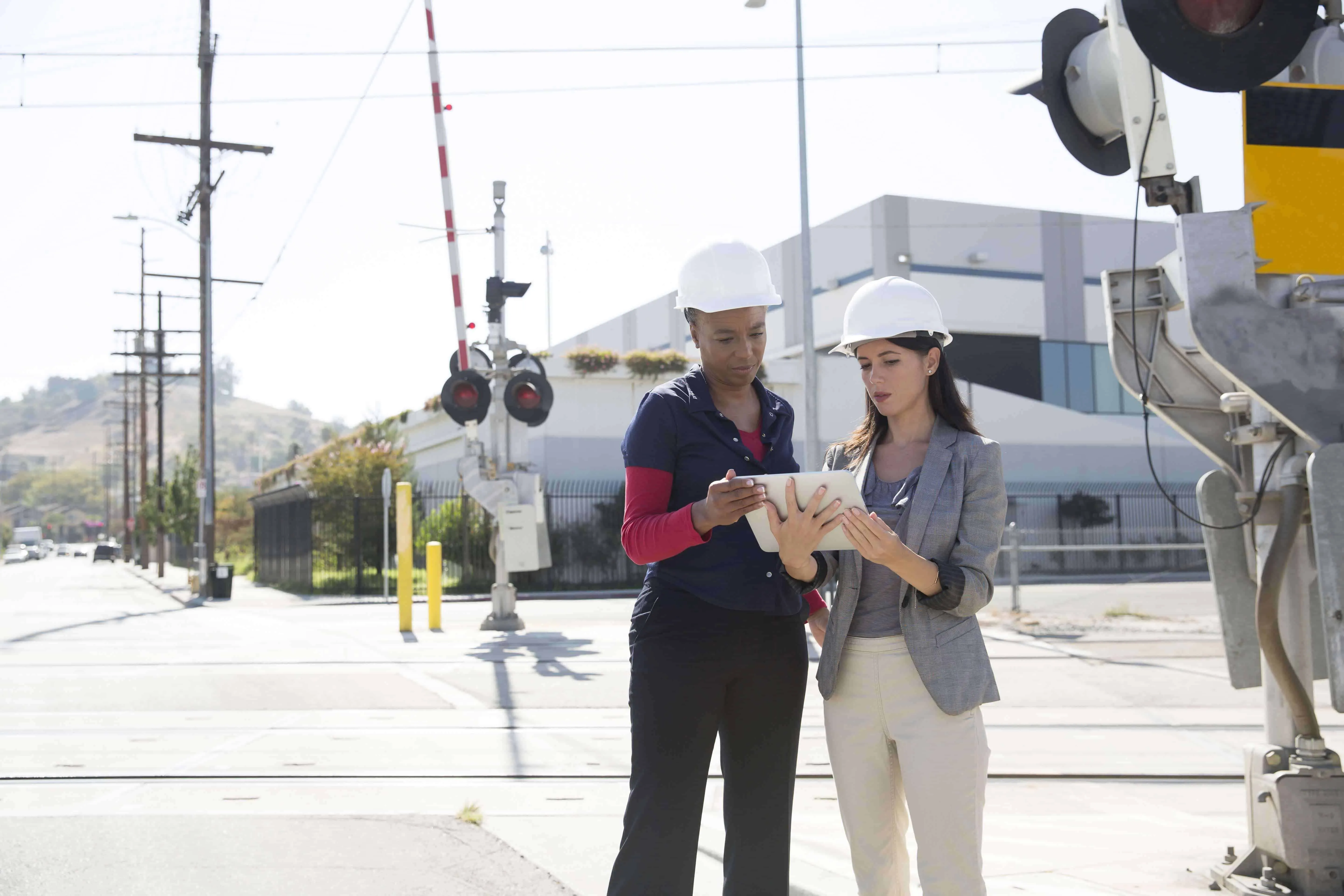 Two engineers in hard hats reviewing plans on a tablet at a railroad crossing