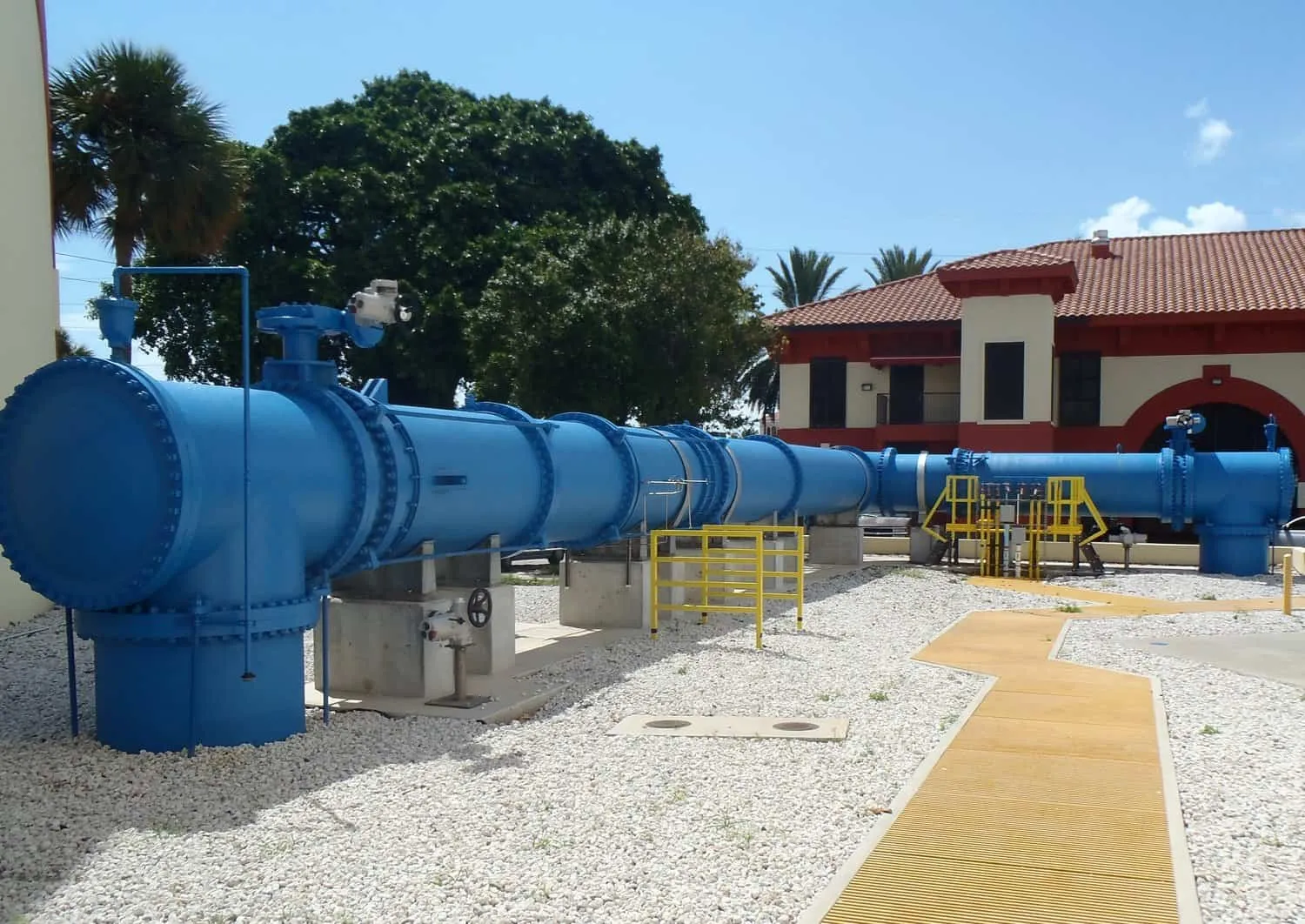 Large blue water pipe and valve assembly outdoors near a tiled-roof building and palm trees